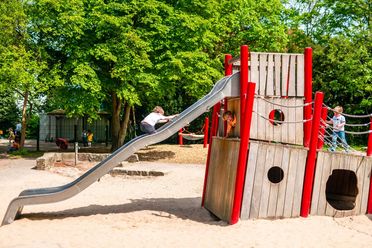 Kinder spielen auf einem Spielplatz im Französischen Garten Celle: Ein Junge rutscht eine Edelstahlrutsche hinunter, während zwei weitere Kinder an einem Holz-Klettergerüst mit roten Pfosten klettern, umgeben von Sand und grünen Bäumen