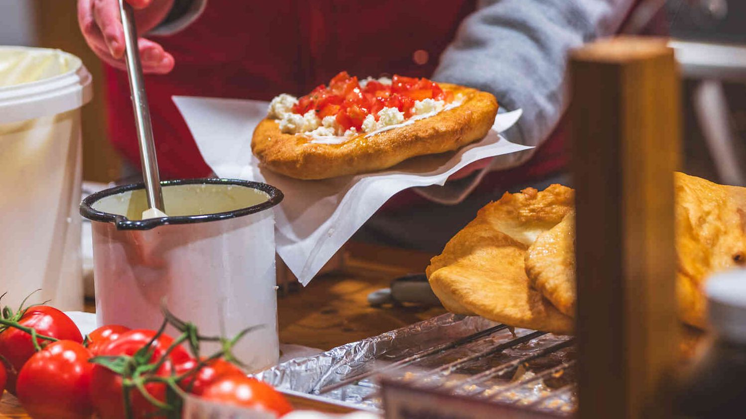 Eine Person belegt ein frisch gebackenes Langos mit Käse und Tomatenwürfeln an einem Marktstand.