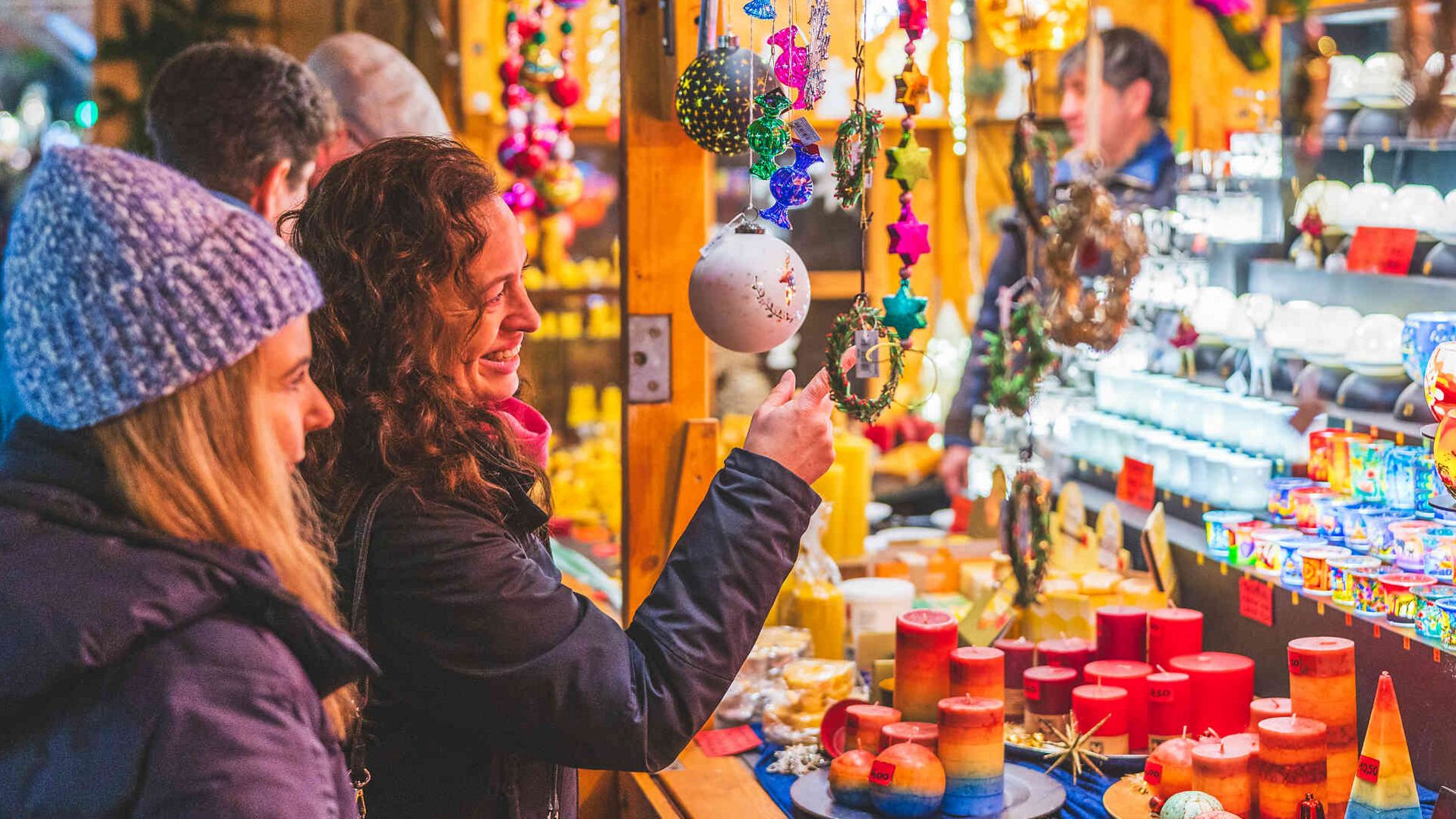 Zwei Frauen betrachten lächelnd bunten Christbaumschmuck an einem Weihnachtsmarktstand. Auf dem Verkaufstisch stehen viele bunte Kerzen. Im Hintergrund liegen Kränze und Keramikwaren aus.
