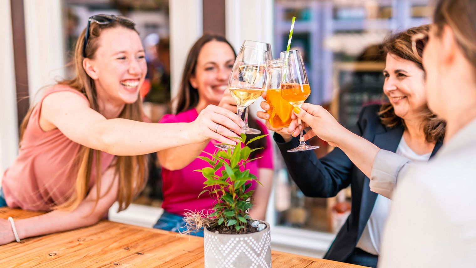 Vier lachende Freundinnen stoßen mit Gläsern in den Händen an einem Holztisch an, umgeben von einer warmen und einladenden Atmosphäre.