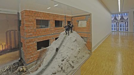 Kunstwerk eines detaillierten Baustellenmodells in einer Ausstellung im Kunstmuseum Celle. Das Modell zeigt ein teilweise zerstörtes Ziegelgebäude mit einem Schutthaufen, auf dem zwei Miniaturfiguren in Anzügen platziert sind. Im Hintergrund sind weitere Gebäude zu sehen, darunter ein hohes Wohngebäude. Der Ausstellungsraum hat einen glatten Holzboden und helle Wände.