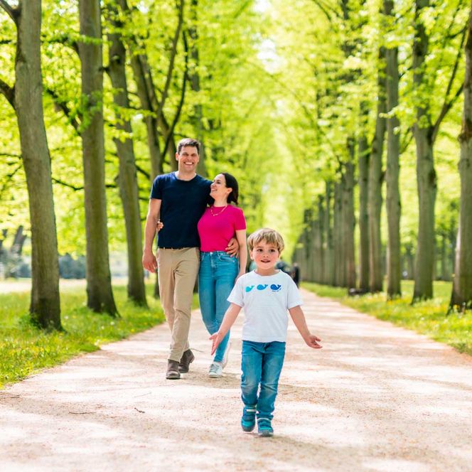 Familie im Französischen Garten Celle: Eltern und Kind laufen lächelnd auf einem sandigen Weg, umgeben von grünen Baumalleen und sonnigem Licht.