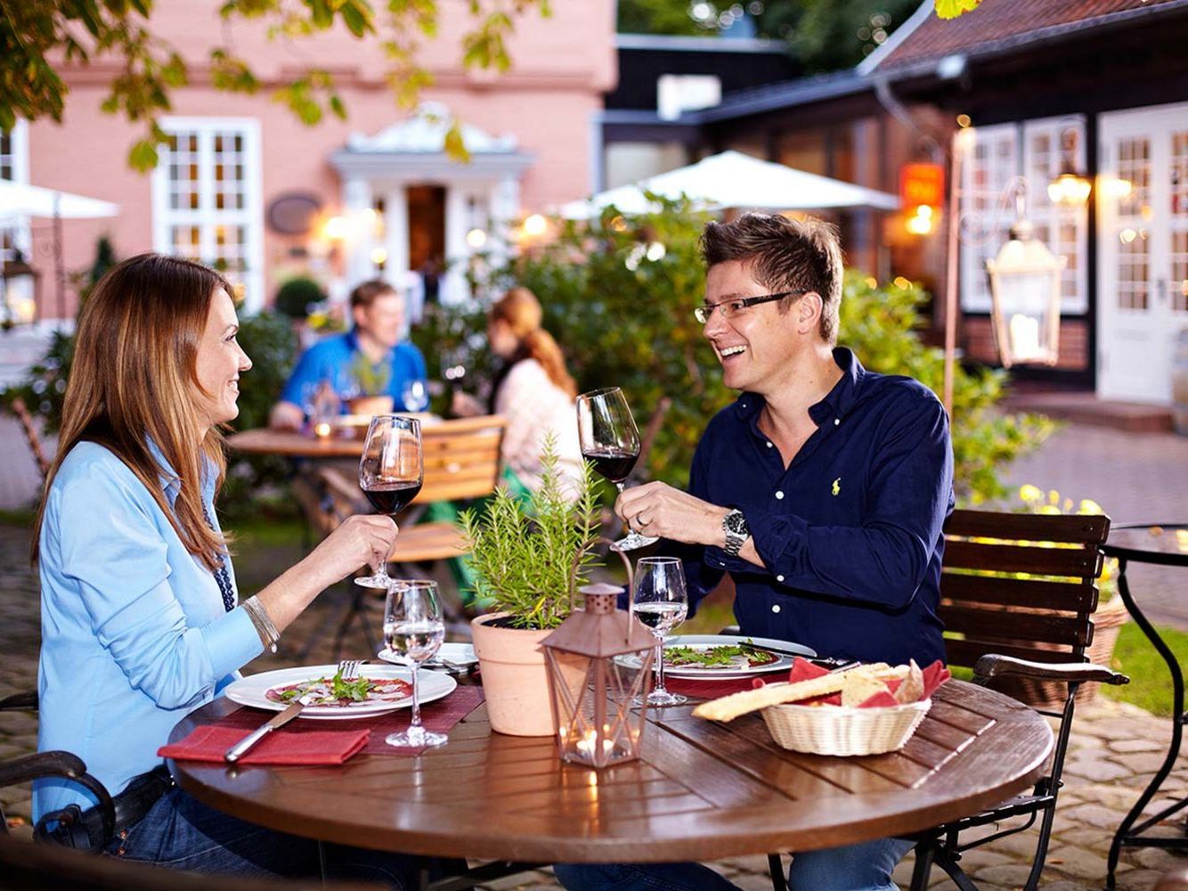 Ein Paar sitzt an einem Holztisch im Freien, hebt die Weingläser und lächelt sich an, während sie ein romantisches Abendessen in einer gemütlichen Atmosphäre genießen.