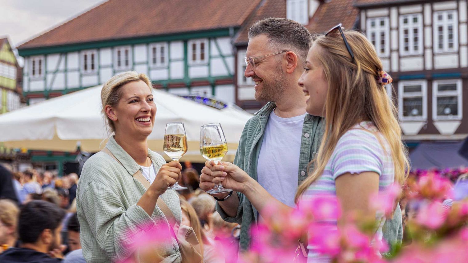 Freundliche Menschen beim Celler Weinmarkt mit Weingläsern, umgeben von blühenden Blumen und traditionellen Fachwerkhäusern
